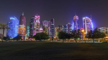 Geceleri Doha 'nın ufuk çizgisi. Yıldızlı gökyüzü Park Timelapse hiper hızından görünüyor, Katar. Ön planda ağaçlar. Aydınlatılmış gökdelenler ve kuleler