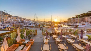 Beautiful sunrise over the harbor in Monaco timelapse. Luxury yachts are illuminated in the morning light. Colorful sky at summer day. Tables and chairs on foreground clipart