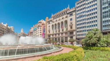 Panorama, Valencia, İspanya 'da Belediye Meydanı Plaza del Ayuntamiento zaman kapsülünü gösteriyor. Büyük mimari, çiçek açan yeşillik ve mavi gökyüzünün altında su jetleri. İnsanlar meydanda geziniyor.