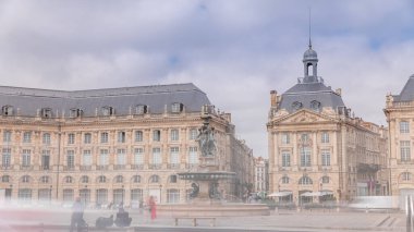 Place de la Bourse zaman atlaması Fransa 'nın Bordeaux şehrinde, Bulutlu mavi gökyüzünün altında klasik mimari ile çevrili Üç Zarafet Çeşmesi ile birlikte. İnsanlar ana meydanda bir tramvay durağında