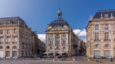 Place de la Bourse, Fransa 'nın Bordeaux şehrinde, bulutlu mavi gökyüzü altında klasik mimari ile çevrili restoranları olan bir yer. Meydanda ve ana caddelerde yürüyen insanlar