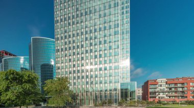 Panorama modern ofis binalarının zaman çizelgesini gösteriyor. Parktaki yeşil ağaçlar tarafından çerçevelenmiş parlak mavi gökyüzü üzerinde yansımaları ve röfleleri olan cam gökdelenler. Brüksel 'de güneş ışığı ve geçen bulutlar