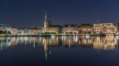 Binnenalster 'in Hamburg, Almanya' daki Belediye Binası ve Nikolai Kilisesi 'ndeki gece programı. Aydınlatılmış tarihi binalar yıldızlı bir gökyüzünün altındaki Alster Gölü sularına yansıyor. Manzaralı şehir manzarası