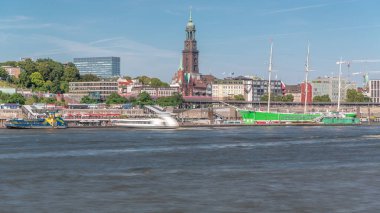 Elbe nehir kenarındaki panoramik zaman çizelgesi ve Almanya 'nın Hamburg kentindeki St. Michael' s Church kulesi. İskeleye yakın teknelerle manzara, Jan Fedder gezinti güvertesi ve oradan geçen feribotlar. Nehir kenarındaki canlı şehir manzarası
