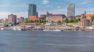 Hamburg limanına giriş ve çıkış yapan bir feribotun zaman çizelgesi. İskelede yolcuları var ve merdivenlerde ilerliyor. Arkaplanda Elbe nehri boyunca tarihi ve modern binalar var. Almanya