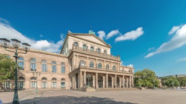 Hanover Devlet Operası 'nın (Staatsoper Hannover) ön görüntüsü. Şehir meydanında canlı mavi bulutlu bir zaman dilimi altında yürüyen insanların olduğu klasik tarzda bir tiyatro. Almanya