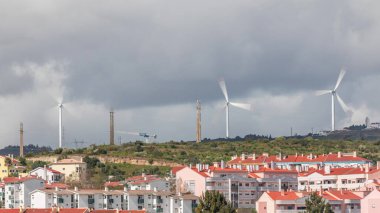 Yenilenebilir Elektrik Enerjisi Zaman Kaynağı Tepedeki Yel Değirmenleri, Rüzgar Çiftliği Hizmetleri. Altında kırmızı çatılar ve arkasında bulutlar olan evler. Yeşil Elektrik Jenerasyonu. Lizbon, Portekiz