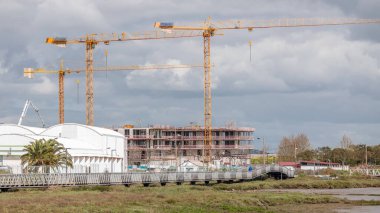 Bulutlu bir zaman dilimi olan bir bina kompleksi üzerinde çalışan birkaç vinç de dahil olmak üzere büyük bir inşaat alanı. Nehir kenarında rıhtımda yürüyen bir köprü var. Lizbon, Portekiz