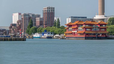 Euromast gözlem kulesi yakınlarındaki Rotterdam şehir manzarası zaman çizelgesi ve Nieuwe Maas Nehri 'ndeki bir turist teknesi. Rıhtımda modern gökdelenler, yüzen bir Çin restoranı ve manzaralı bir liman manzarası yer alıyor.