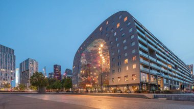 Rotterdamse Markthal 'ın dış görünüşünün geceden güne geçişi. Dükkanları ve restoranları olan ikonik bir bina. Meydanda gezinen insanlar. Gökdelenler ve arka planda canlı bir şehir manzarası