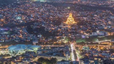 Aydınlanmış Tsminda Sameba Katedrali ya da Tiflis 'in Kutsal Üçlü Katedrali ve Kamu Hizmetleri Salonu hava gününden gece geçiş zamanına. Tiflis, Gürcistan 'daki ana katedral. Arka plandaki evler