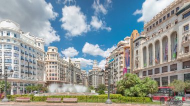 Valencia, İspanya 'da Belediye Meydanı' ndaki (Plaza del Ayuntamiento Meydanı) zaman atlaması. Büyük mimari, çiçek açan yeşillik ve bulutlu bir gökyüzünün altında su jetleri. İnsanlar canlı meydanda geziniyor..