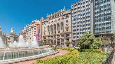Panorama, İspanya 'nın Valencia şehrinde Belediye Meydanı' nda (Plaza del Ayuntamiento Meydanı) fıskiyeyi gösteriyor. Büyük mimari, çiçek açan yeşillik ve mavi gökyüzünün altında su jetleri. İnsanlar meydanda geziniyor.