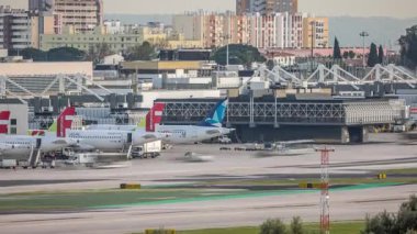 Lizbon Havaalanı Humberto Delgado saatine park etmiş uçaklar hareket ediyor. Pist aktivitesi, terminaller, şehir silueti ve açık bir gökyüzü altında hareket halindeki farklı havayolu şirketlerinin uçakları. Lizbon, Portekiz.