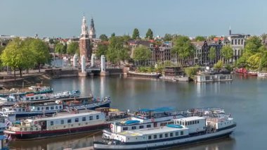 Amsterdam 'ın şehir panoramasının önünde Montelbaanstoren kulesi ve kanal boyunca uzanan şehir manzarası yer almaktadır. Eski kentin rıhtımında tarihi Hollanda evleri, mavnalar ve yüzen evler