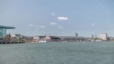 Amsterdam Merkez İstasyonu 'nun kıvrımlı çatısı ve Amstel Nehri zaman dilimi ile panoramik manzarası. Bir feribot, bulutların hareketli olduğu güneşli bir gökyüzünün altında nehri geçer. Amsterdam, Hollanda