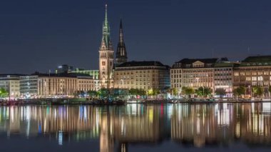 Binnenalster 'in Hamburg, Almanya' daki Belediye Binası ve Nikolai Kilisesi 'ndeki gece programı. Aydınlatılmış tarihi binalar yıldızlı bir gökyüzünün altındaki Alster Gölü sularına yansıyor. Manzaralı şehir manzarası