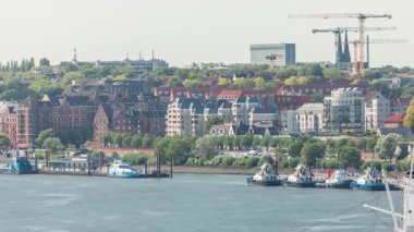 Almanya 'nın Hamburg kentinde Elbe nehir kenarının hava zaman çizelgesi. İskeleye yakın tekneler, marina ve geçen gemilerle manzara. İnşaat alanında vinçleri olan evler. Elbe Nehri kıyısındaki canlı şehir manzarası
