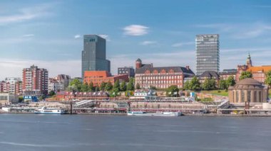 Hamburg limanına giriş ve çıkış yapan bir feribotun zaman çizelgesi. İskelede yolcuları var ve merdivenlerde ilerliyor. Arkaplanda Elbe nehri boyunca tarihi ve modern binalar var. Almanya