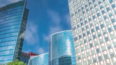 Modern ofis binalarının zaman çizelgesi. Parktaki yeşil ağaçlar tarafından çerçevelenmiş parlak mavi gökyüzü üzerinde yansımaları ve röfleleri olan cam gökdelenler. Brüksel, Belçika 'da güneş ışığı ve geçen bulutlar