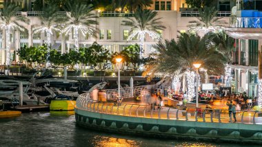 Yürüyen insanlarla dolaş ve Dubai Marina Kanalı üzerinde trafiği olan köprü gece vakti, BAE. Palmiyeler, tekneler ve kulelerle rıhtımın manzarası. Yapay kanallı bölge