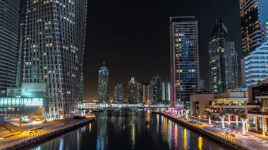 Gece Dubai Marina 'da gezinti ve kanal saatleri, BAE. Palmiyeler, yüzen tekneler ve yanan kulelerle köprüden hava manzarası. Yatı olan liman