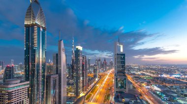 Dubai şehir mimarisi günden geceye geçiş zamanı. Şeyh Zayed yolu üzerindeki aydınlık gökdelenler ve trafik manzaralı üst hava manzarası. Güzel bulutlu gökyüzü