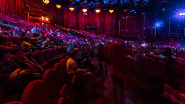Spectators gather in the auditorium and watch the show in theatre timelapse. Large hall with red armchairs seats. Viewers filling places until turn off the light. Panoramic view from left side