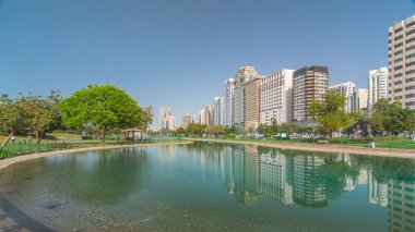 Abu Dabi 'nin sahil şeridi boyunca uzanan Corniche Bulvarı plajı parkında arka planda gökdelenler var. Lake Park 'tan yansıtmalı görüntü. Bir yanda ağaçlar