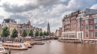 Amstel Nehri, rıhtım ve Munttoren kulesinin yer aldığı Amsterdam şehir merkezinin panoramik zaman atlaması. Tekneler bulutlu gökyüzünün altında ileri geri yüzerler. Köprüden manzaralı şehir manzarası. Hollanda