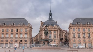Place de la Bourse zaman atlaması Fransa 'nın Bordeaux şehrinde, Bulutlu mavi gökyüzünün altında klasik mimari ile çevrili Üç Zarafet Çeşmesi ile birlikte. İnsanlar ana meydanda bir tramvay durağında