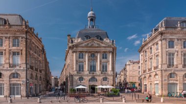Place de la Bourse, Fransa 'nın Bordeaux şehrinde, bulutlu mavi gökyüzü altında klasik mimari ile çevrili restoranları olan bir yer. Meydanda ve ana caddelerde yürüyen insanlar