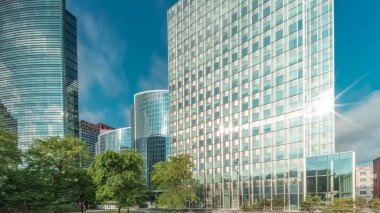 Panorama modern ofis binalarının zaman çizelgesini gösteriyor. Parktaki yeşil ağaçlar tarafından çerçevelenmiş parlak mavi gökyüzü üzerinde yansımaları ve röfleleri olan cam gökdelenler. Brüksel 'de güneş ışığı ve geçen bulutlar