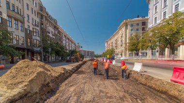 Turuncu üniformalı işçiler yol inşaatında toprak kazıyor ve zaman aşımına uğruyor. Arka planda endüstriyel sarı kazıcı. Kavşaktaki tramvay raylarının yeniden yapılandırılmasının görüntüsü
