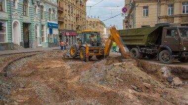 Endüstriyel sarı kazıcı toprağı taşıyor ve yol inşaatındaki bir çöp kamyonuna yüklüyor. Kavşaktaki tramvay raylarının yeniden yapılandırılmasının görüntüsü