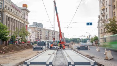 Yol inşaatı alanındaki panoramik zaman dilimine vinçle beton plakalar yerleştiriliyor. Şapkalı ve üniformalı endüstriyel işçiler. Şehir caddesindeki tramvay raylarının yeniden inşası