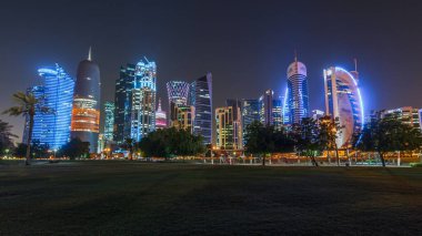 Geceleri Doha 'nın ufuk çizgisi. Yıldızlı gökyüzü Park Timelapse hiper hızından görünüyor, Katar. Ön planda ağaçlar. Aydınlatılmış gökdelenler ve kuleler