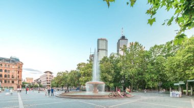 Frankfurt, Almanya merkezindeki Opera Meydanı (Opera Meydanı) zaman atlaması, arka planda gökdelenler ve kulelerle çevrili ve akşam gökyüzü altında ağaçlarla çevrili bir çeşme ve şehir hayatı sergiliyor.
