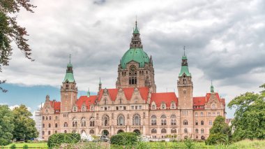 Hanover 'in Yeni Belediye Binası' nın hiperlapesi. Bulutlu bir gökyüzünün altında park ve göl ile çevrili tarihi bir kale gibi bir yapı. Su ve yeşillik üzerindeki yansımalar bu ikonik şehir manzarası zaman çizelgesini oluşturuyor. Almanya