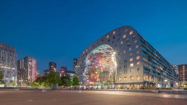 Rotterdamse Markthal 'ın dış görünüşünün geceden güne geçişi. Dükkanları ve restoranları olan ikonik bir bina. Meydanda gezinen insanlar. Gökdelenler ve arka planda canlı bir şehir manzarası