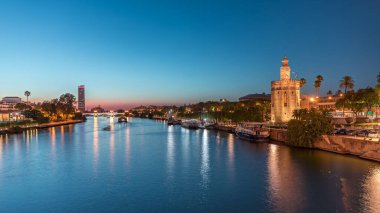 İspanya, Seville 'deki Torre del Oro gözlem kulesi yakınlarındaki Guadalquivir Nehri boyunca yüzen bir tur teknesi. Günbegün geçiş zamanı, şehrin ışıkları su ve deniz manzarasını yansıtıyor.