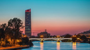 Guadalquivir Nehri yakınlarındaki Torre Sevilla gökdeleninin yer aldığı Seville 'in ufuk çizgisi gece gündüz geçiş zamanı. Aydınlatılmış Triana Köprüsü su, modern ve geleneksel mimariyi yansıtıyor