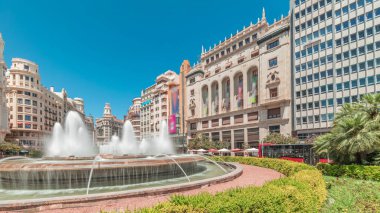 Panorama, İspanya 'nın Valencia şehrinde Belediye Meydanı' nda (Plaza del Ayuntamiento Meydanı) fıskiyeyi gösteriyor. Büyük mimari, çiçek açan yeşillik ve mavi gökyüzünün altında su jetleri. İnsanlar meydanda geziniyor.