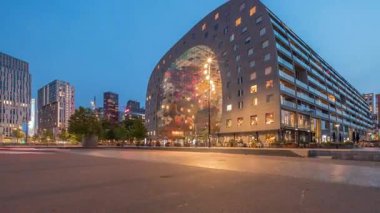 Rotterdamse Markthalleri 'nin dış görünüşü günden geceye değişiyor. Dükkanları ve restoranları olan ikonik bir bina. Meydanda gezinen insanlar. Gökdelenler ve arka planda canlı bir şehir manzarası
