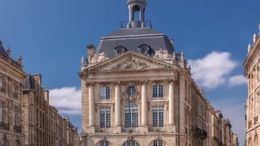 Place de la Bourse, Fransa 'nın Bordeaux şehrinde, bulutlu mavi gökyüzü altında klasik mimari ile çevrili restoranları olan bir yer. Meydanda ve ana caddelerde yürüyen insanlar