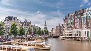Amstel Nehri, rıhtım ve Munttoren kulesinin yer aldığı Amsterdam şehir merkezinin panoramik zaman atlaması. Tekneler bulutlu gökyüzünün altında ileri geri yüzerler. Köprüden manzaralı şehir manzarası. Hollanda