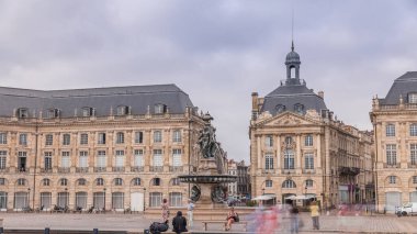 Place de la Bourse zaman atlaması Fransa 'nın Bordeaux şehrinde, Bulutlu mavi gökyüzünün altında klasik mimari ile çevrili Üç Zarafet Çeşmesi ile birlikte. İnsanlar ana meydanda bir tramvay durağında