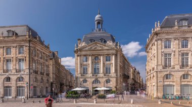 Place de la Bourse, Fransa 'nın Bordeaux şehrinde, bulutlu mavi gökyüzü altında klasik mimari ile çevrili restoranları olan bir yer. Meydanda ve ana caddelerde yürüyen insanlar