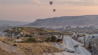 İnsanlar, Türkiye 'nin Kapadokya kentindeki olağandışı kayalık hava zaman çizelgesinin üzerinde açık bir sabah gökyüzünde renkli sıcak hava balonlarıyla güneşin doğuşunu seyrediyorlar.