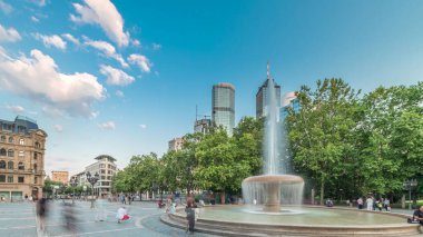 Frankfurt, Almanya merkezindeki Opera Meydanı (Opera Meydanı) zaman atlaması, arka planda gökdelenler ve kulelerle çevrili ve akşam gökyüzü altında ağaçlarla çevrili bir çeşme ve şehir hayatı sergiliyor.